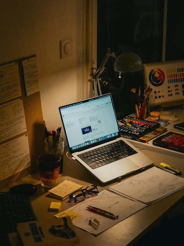 A desk at night with a laptop, books and loose papers